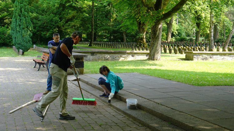 Teilnehmende am Kriegsgräber-Pflegeeinsatz reinigen Pflasterfugen und fegen mit einem Besen.
