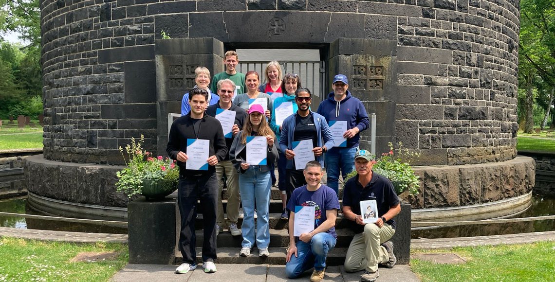Teilnehmende am Kriegsgräber-Pflegeeinsatz halten die Dankurkunden des Volksbunds in den Händen und stehen vor dem Kriegstoten-Denkmal auf dem Frankfurter Hauptfriedhof.