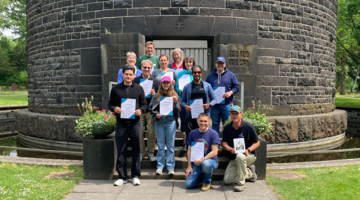 Teilnehmende am Kriegsgräber-Pflegeeinsatz halten die Dankurkunden des Volksbunds in den Händen und stehen vor dem Kriegstoten-Denkmal auf dem Frankfurter Hauptfriedhof.
