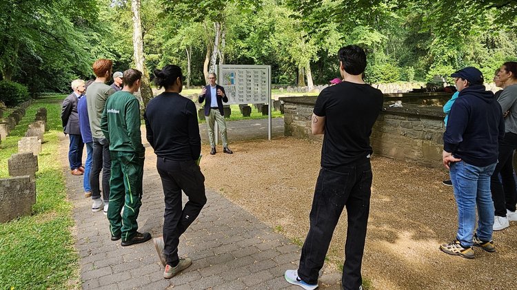 Teilnehmende am Kriegsgräber-Pflegeeinsatz hören dem Bericht von Dr. Götz Hartmann über die Geschichte der Kriegsgräber auf dem Frankfurter Hauptfriedhof zu.